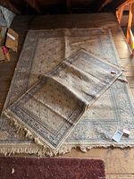 Photo showing two beige and blue wool rugs, one large and one smaller, laid on wooden floor with visible damage and fringe edges.