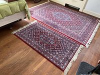 Photo showing two rectangular wool Oriental rugs with matching medallion patterns and fringed edges laid on hardwood floor near bed.