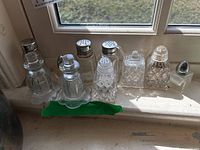 Seven vintage-style glass salt and pepper shakers placed on a window ledge, showing variety in shapes and metal tops.