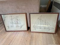 Two framed ship prints shown leaning on floor. Both frames are wood with glass. Prints are detailed ship drawings in black ink on beige paper.