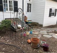 Overall view of the black shepherd hooks with colorful solar lanterns, plant pots, and decorative spheres placed on soil ground near house exterior.