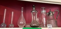 Seven glass and metal items on a red backdrop - two glass bud vases, two decanters, glass bowl, pewter vase, glass pitcher with silver plated top