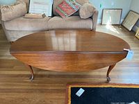 Wooden oval drop leaf coffee table closed position showing top and legs with floor and sofa in background.
