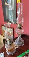 Two vintage oil lamps placed on a table against a red wall. One lamp has a beaded glass chimney and a handle on the glass base, the other has a taller smooth chimney without a handle.