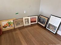Six framed vintage prints lined against a wall on wood floor, including fruit and scenic artwork. Visible damage on some frames and prints, some water staining.