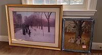 Two framed and matted prints displayed side-by-side on floor by window, showing a winter park scene and children playing around a tree.