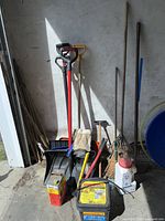 Image showing plastic shovels, rakes, Roofmelt container, Ortho Triox vegetation killer bottle, windshield scrapers, and edger tools arranged upright against a wall.