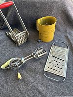 Photo showing the french fry press, cylindrical flour sifter, handheld grater, and manual blender/beater with yellow handle.