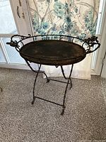 Full view of the antique fold-up metal tea table showing the round tray top with decorative handles and foldable legs.