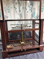 Front view of antique brass scale mounted in original wooden and glass case. Brass balance beam and central pillar visible, scale is missing parts the beam rests on the base inside the case.
