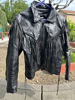 Front view of black leather fringe jacket hanging outdoors, showing front zipper, collar, and chest fringe details.