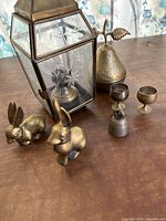 Photo showing all items in the lot including hurricane lamp, brass bunnies, brass pear container, goblets, and pepper shaker arranged on a brown surface.