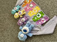 Group photo of four vintage Care Bears plush toys arranged on top of a light purple Care Bears women's XL half top shirt with a colorful graphic patch of Care Bears.