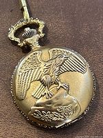 Close-up of the gold tone pocket watch cover with embossed eagle design perched on a rock with floral accents.