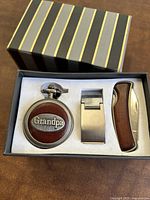 Box set with pocket watch, money clip, and folding pocket knife displayed in foam insert inside striped box.