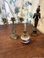 Photo showing the three brass items: candelabra, oil lamp, and incense holder arranged on a wooden surface with floral curtain background.