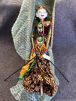Front view of the Indonesian Golek rod puppet showing detailed painted face, wooden articulated arms, and colorful traditional batik dress with green and yellow ribbons. Puppet is placed on bubble wrap.