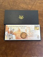 Black presentation box with gold emblem containing a certificate card featuring Pope John Paul II image, Vatican date stamp, and embedded coin in protective case.