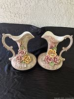 Front view of two large porcelain pitcher-style vases with floral decorations and ornate handles.