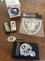 Photo showing two Raiders logo watches, reflective decal in packaging, Raiders logo keychain, wallet with Raiders helmet design, and boxed glass Christmas ornament.