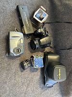 Photo showing three cameras, one lens attachment, vintage slide reader, and Minolta soft case arranged on fabric surface.