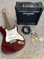 Full lot of red Hamer Slammer electric guitar, black Behringer amplifier, cable, and guitar picks shown on carpeted floor.