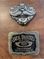 Pair of vintage metal belt buckles, one 'Mustache Rides 5¢' and one 'Jack Daniels Old Time Tennessee Whiskey'.
