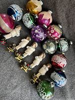 Twelve vintage glass Christmas ball ornaments in varied colors and white decorative paint, alongside five clip-on stem ornaments featuring small felt figures dressed in cream winter clothing and mounted on gold metal clips decorated with pinecones and berries.