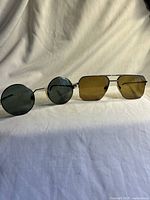 Both pairs side by side on a draped white surface showing round green tinted and rectangular brown tinted lenses