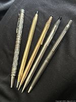 Five vintage pens and mechanical pencils laid flat on black cloth, showing variety in metal finishes and designs.