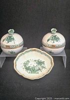 Front view of two covered candy dishes with floral lids and one scalloped small plate with green floral basket pattern, arranged on black background.