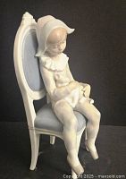 Full side view of the porcelain figurine showing the child figure seated on a blue cushioned ornate white chair in Harlequin costume.