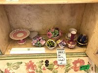 Display of all porcelain items in a small cabinet shelf showing multiple small vases, trinket box, pedestal dish, and flower-shaped pieces.