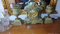 Full view of green stone mantel clock with brass mounts flanked by two urns