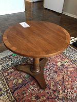 Front angled view of a wooden round pedestal side table on a patterned rug, showing the tabletop and the tripod base.