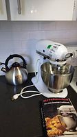 Photo of white KitchenAid stand mixer K45SS with stainless steel bowl and whisk attachment alongside LuxGrace stainless steel kettle, placed on kitchen counter against white tiled backsplash, showing power cord and KitchenAid cookbook.