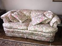 Front angle view of the floral upholstered love seat showing two matching cushions on a wooden floor with area rug nearby.