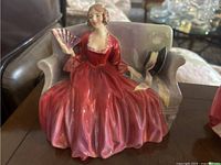 Front view of figurine showing seated woman in red-pink dress holding fan on lavender bench