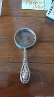 Magnifying glass with detailed floral silver handle and round glass lens shown on wooden surface.