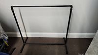 Black metal pipe clothes rack standing against a white wall on a brown carpeted floor, showing the full front view of the rack.