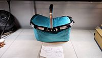 Teal collapsible picnic basket with metal handle placed on tile floor with a fan and cardboard visible in background. Instruction sheet laid in front of basket.