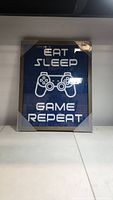 Framed picture with blue background featuring the phrase 'EAT SLEEP GAME REPEAT' and a game controller graphic, displayed on a white surface.