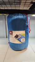Full view of the blue Outbound sleeping bag packed inside its blue storage sack with red drawstrings on a shelf.