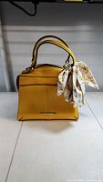 Front view of the mustard yellow structured handbag with single handle and floral scarf tied to it.