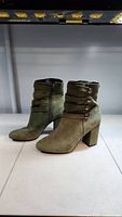 Side profile of olive green suede ankle boots showing decorative straps and block heel.