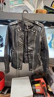 Front view of the women's black leather jacket hanging on a black hanger, showing zippers and snap button details