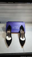 Front view of pair of black suede high heel shoes with Stuart Weitzman box behind