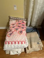 Bundles of various blankets piled on a wooden floor against a wall, showing textures and colors such as pink, cream and striped wool patterns.