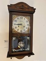 Frontal view of the brown wooden pendulum wall clock showing the clock face, numbers, hands, and decorative gold corners.