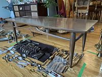 Angle view of the coffee table showing wood grain top, angled tapered legs, and horizontal support bar, placed on hardwood floor with various silverware items arranged underneath.